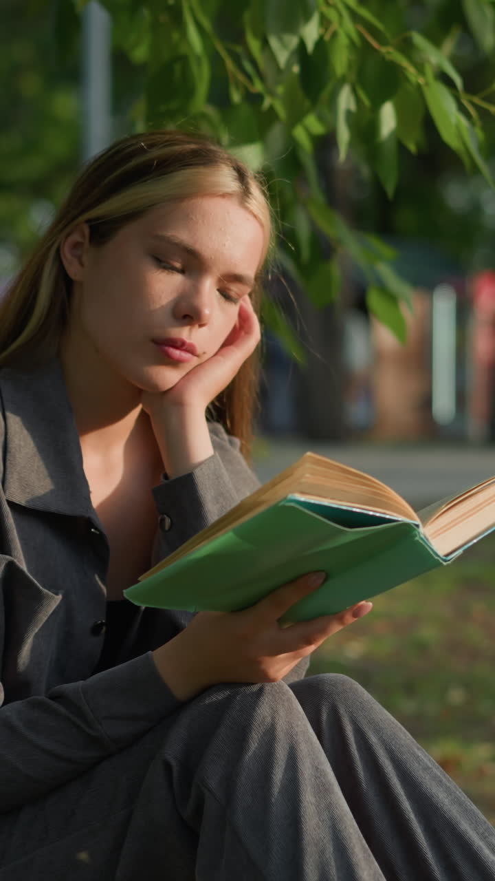 dama sentada bajo un árbol con la cabeza apoyada en la mano derecha, hojeando un libro y apareciendo de mal humor, la luz del sol toca suavemente su cara mientras el fondo suave y borroso presenta objetos lejanos