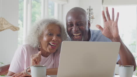 Pareja-Mayor-Sentada-Alrededor-De-Una-Mesa-En-Casa-Haciendo-Videollamadas-En-Una-Computadora-Portátil