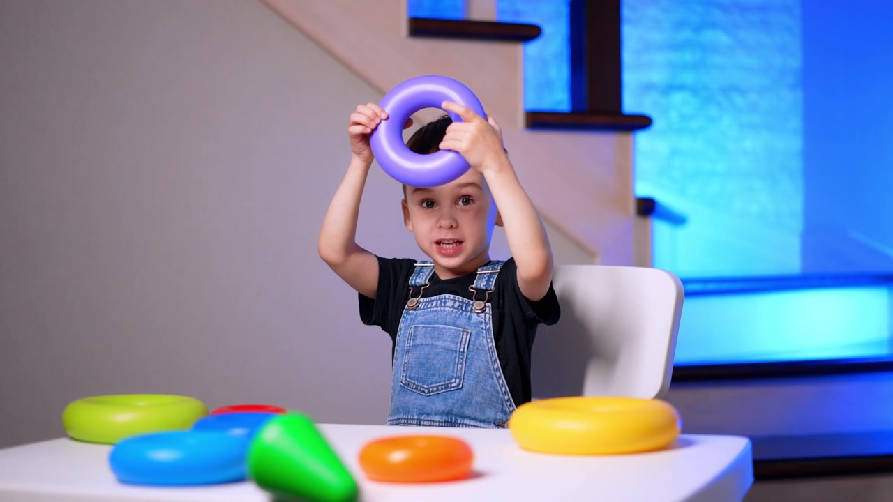 Young boy holding purple ring toy on head at table. A child sits with colorful stacking rings on a white desk indoors.