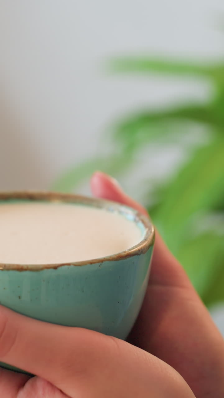 Closeup person sipping latte from teal cup, hands cradling warm foam in ceramic mug, soft sunlight and blurred green plant background, calm cozy atmosphere, mindful slow morning ritual and gentle