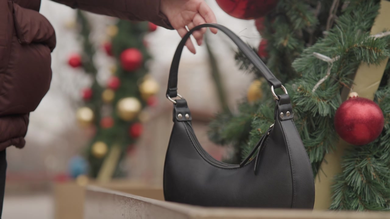 Borsa nera appesa a una ghirlanda, scena di mercatino di strada con luci sfocate dell'albero di Natale, palline rosse e festoni sempreverdi, manica di un acquirente brevemente visibile mentre la borsa scivola su una ringhiera di legno, pelle