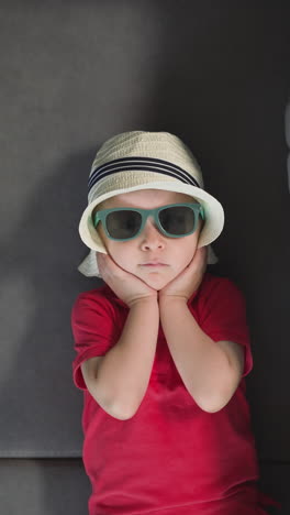 adorable toddler adjusts straw hat lying on soft grey sofa. stylish little boy in sunglasses relaxes after arrival in hotel room upper closeup