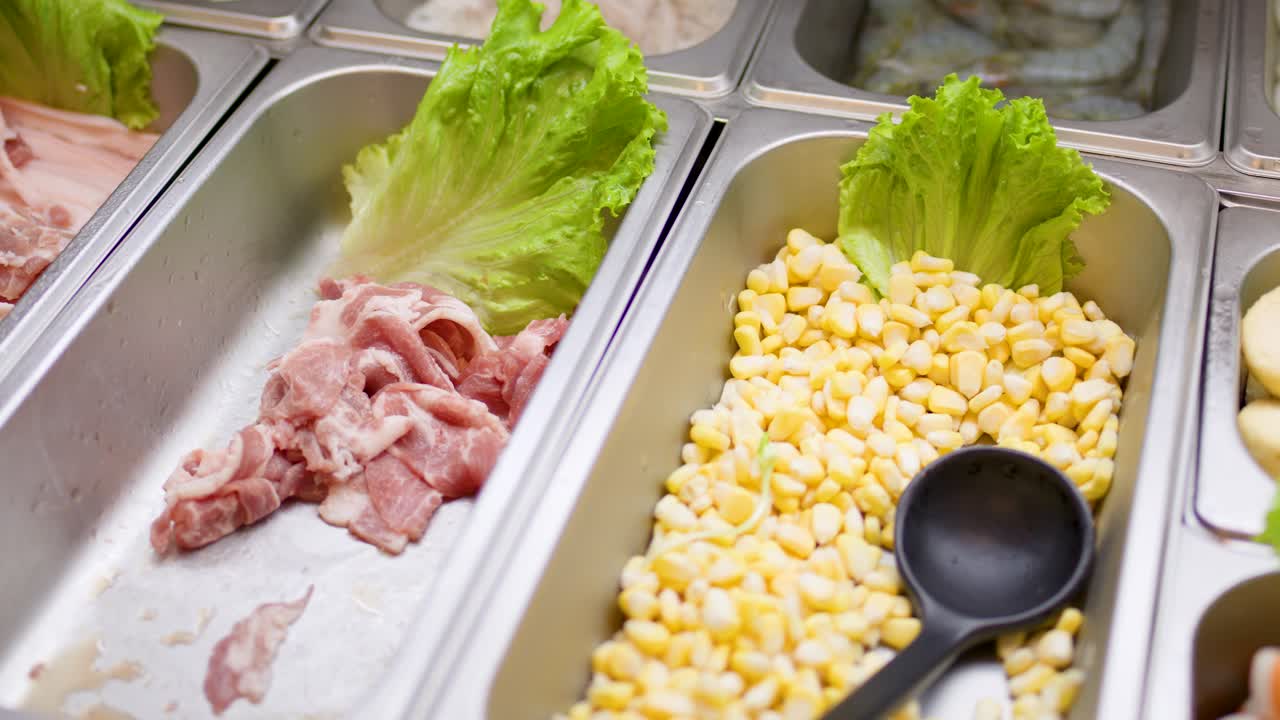 Brightly lit buffet displays various raw hotpot ingredients, vegetables, and noodles in stainless steel trays