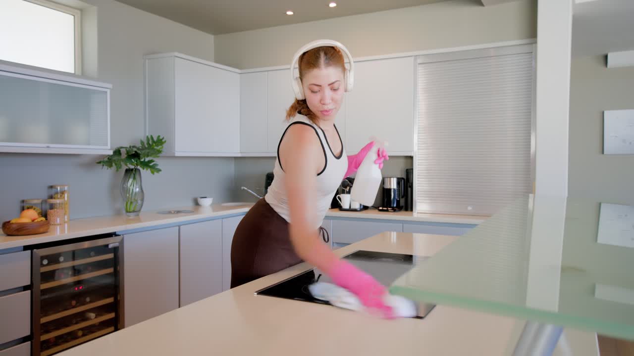 Donna che individua aloni, spruzza da una bottiglia e pulisce con un panno un piano cottura a induzione, indossando guanti rosa.