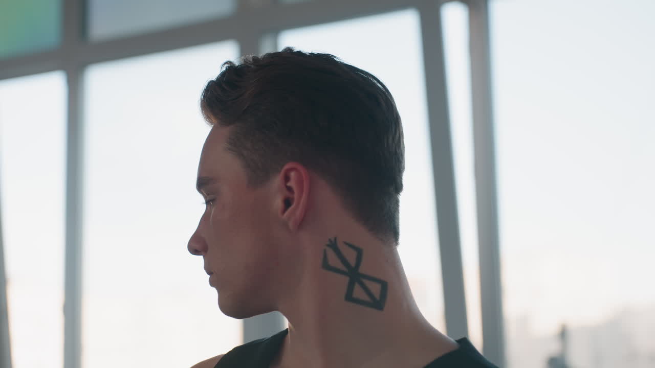 young man removes hat to reveal bold neck tattoo in sunlit studio with tall windows behind subtle gleam on skin, dust motes floating, gentle breeze in hair, elegant dancer portrait with dramatic mood