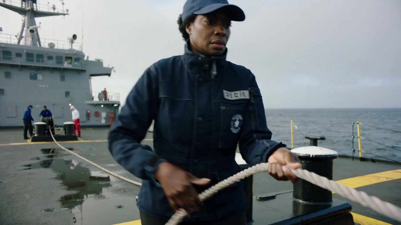 Dæksmand trækker på fordækket i marineblå jakke, mens spillet starter og strammer fortøjningslinen for at sikre skibet