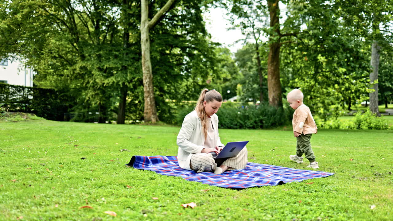 En ung mor sidder på et picnictæppe i en grøn park med sin lille søn. Den lille søn løber ved siden af sin mor og kigger interesseret på den bærbare computers skærm. Den unge mor arbejder fjernstyret.