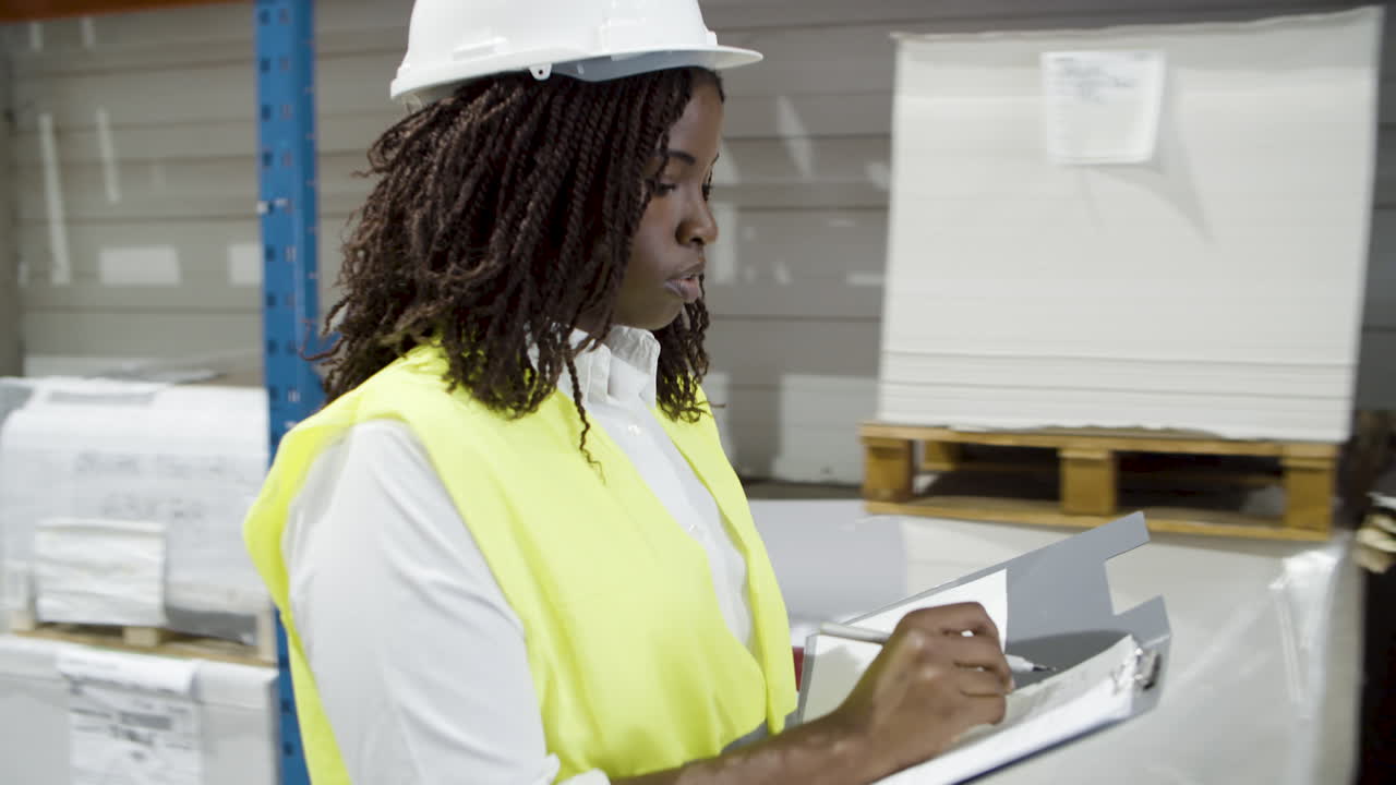 Serious African American female worker in helmet counting goods in stock