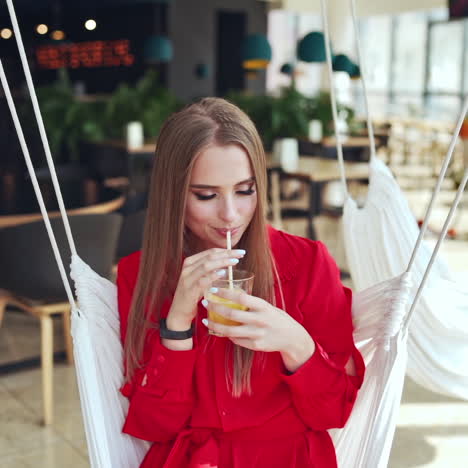 attractive lady in red dress in a cafe. young beautiful woman is drinking juice on a white swing. portrait of a pretty lady relaxing. slow motion
