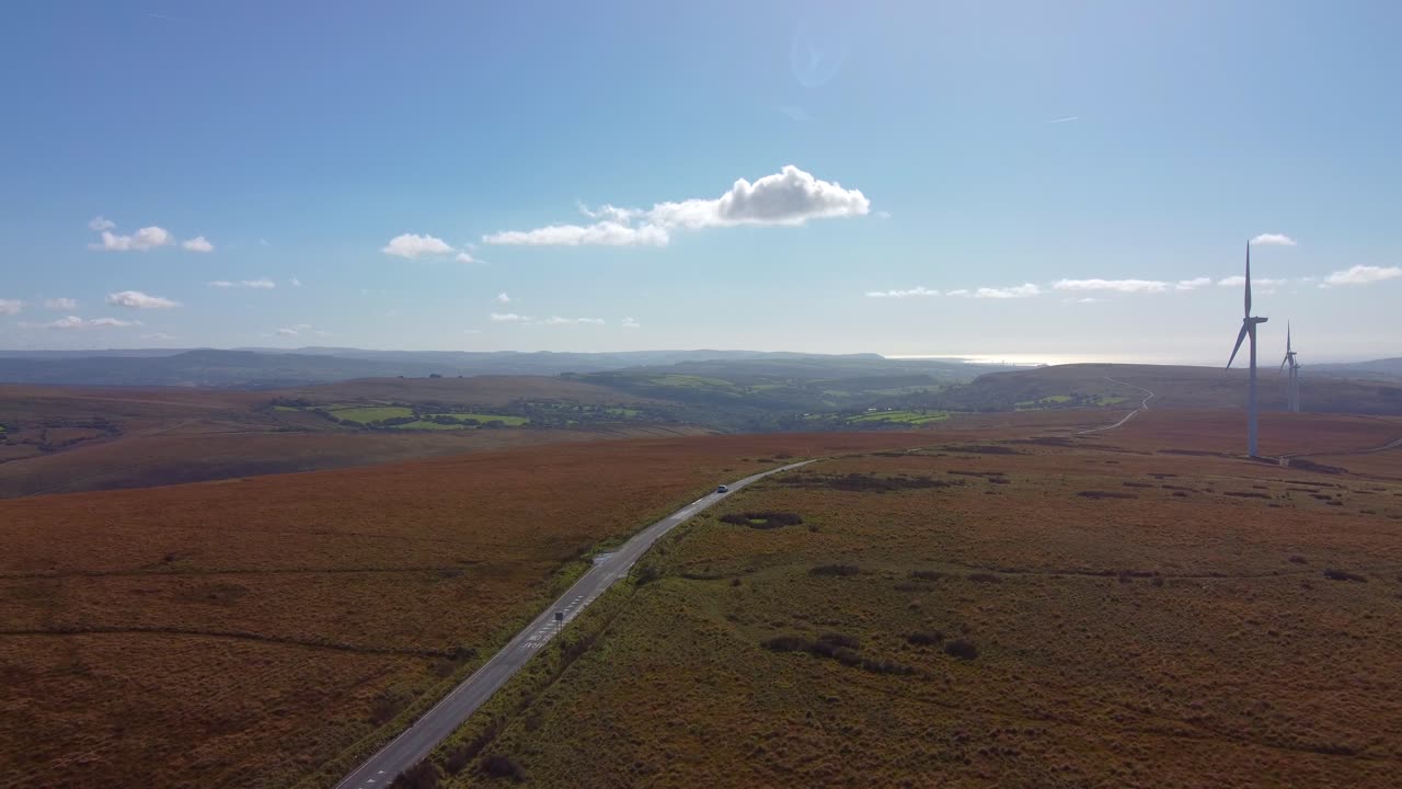 Aerial View of Lone Electric Car Driving Across Long Straight Road on Wind Turbine Farm and Welsh Countryside Moors and Fields 4K