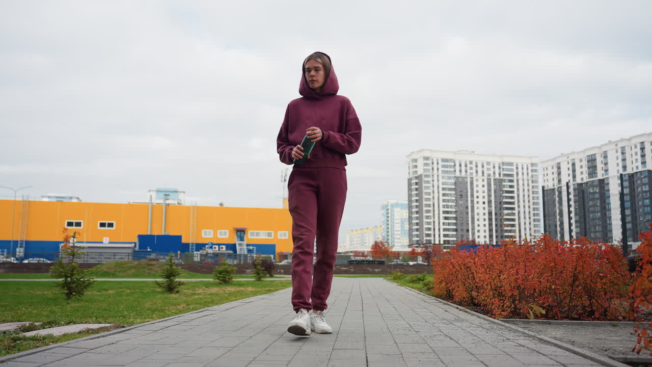 Young sporty woman in maroon hoodie strides along paved city park path with reusable water bottle, embracing energetic lifestyle amid high rise skyline, autumn shrubs, overcast sky, inspiring fitness
