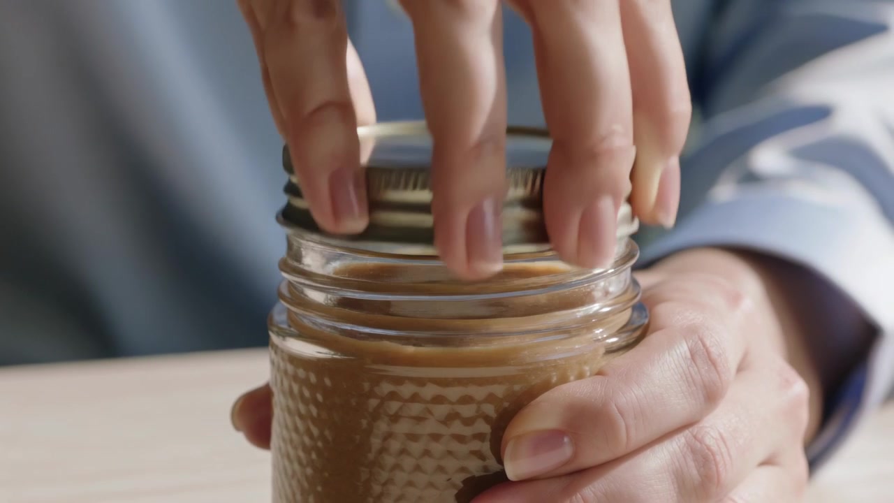 Gripping metal ring, man wearing light blue shirt twisting jar on tabletop, revealing brown spread