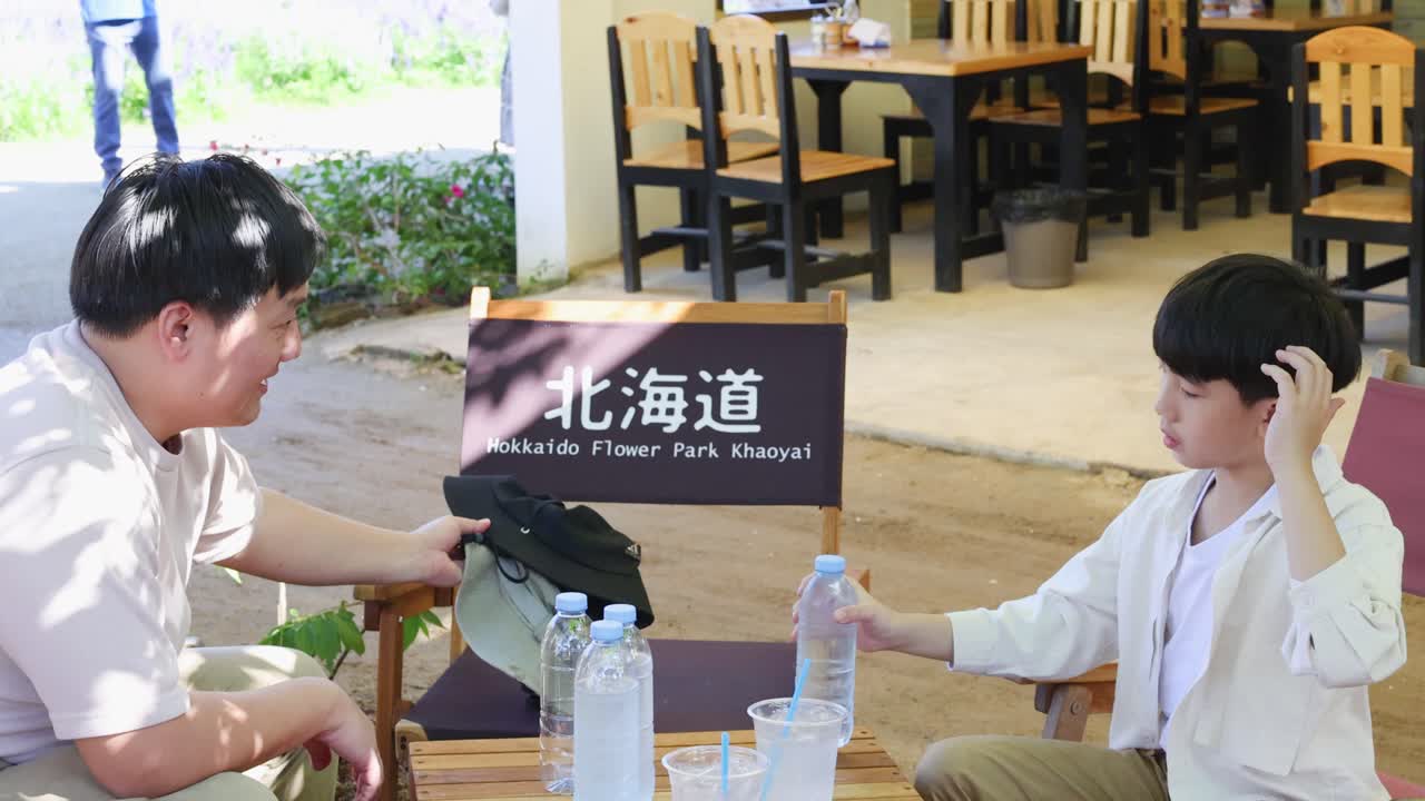 旅行の休憩中、日当たりの良い屋外カフェでリラックスし、ペットボトルの水を飲む父親と息子。