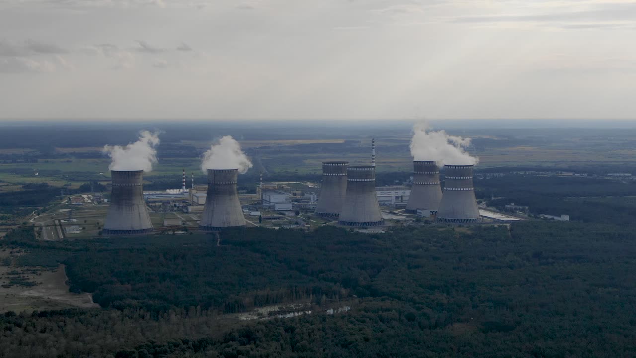 Aerial view of a nuclear power plant with multiple cooling towers emitting steam, surrounded by a forest.