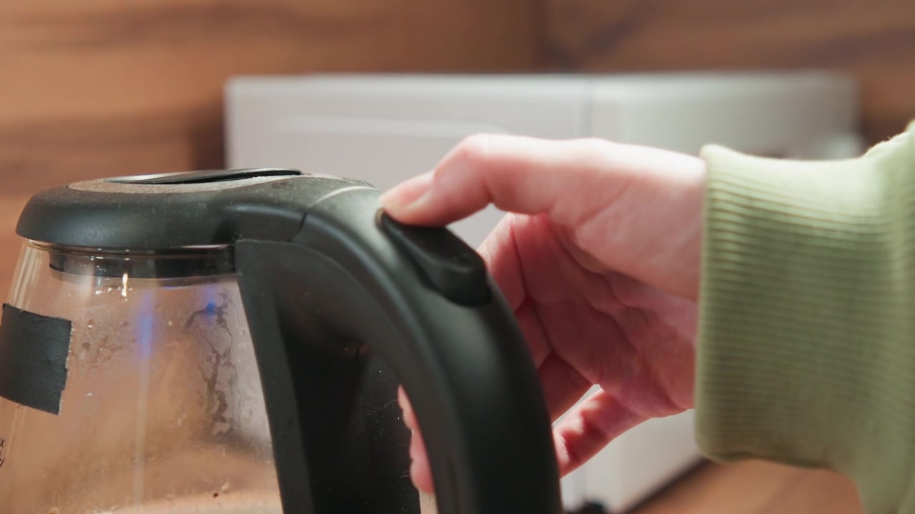 Hand operates kettle control button beside glass body filled with rising steam and condensation in bright home kitchen, creating warm reflections and clear signs of freshly heated water
