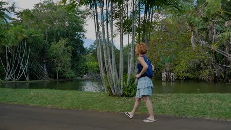 a slow motion side shot folllowing a woman walking near a pond in a garden full of tropical plants