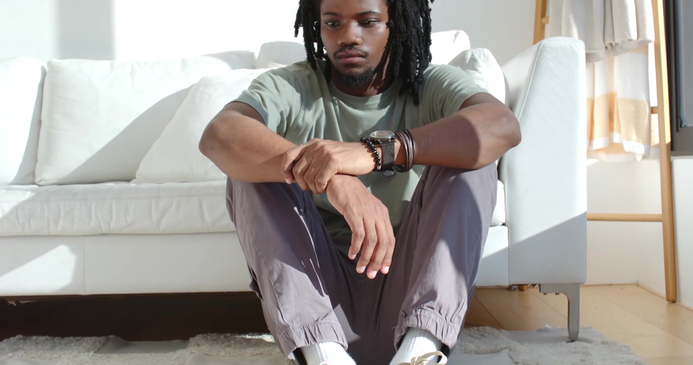Adult African man sitting on rug by white sofa, adjusting wristwatch as sunlight crossing rug