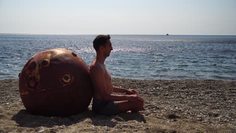 man doing yoga exercise outdoors near to an old rusty floating marine mine on the beach with rocky shore and sea background. healthy lifestyle, pollution, nature protection, war and peace concept