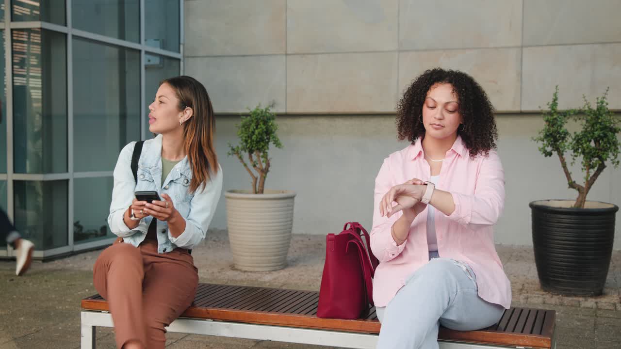 African American women on bench left scrolling phone, right checking watch waiting as people pass