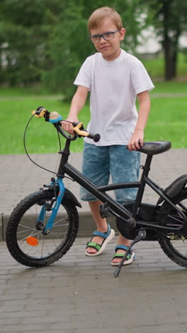 un niño joven con gafas y sandalias camina hacia una bicicleta estacionada con una ligera sonrisa en su rostro, vestido con una camiseta blanca y pantalones cortos
