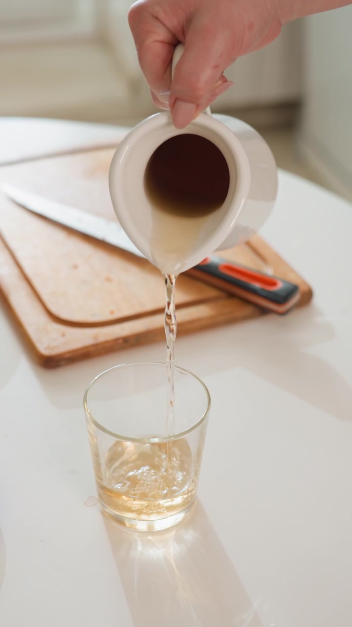 Una brocca di ceramica bianca si inclina su un bicchiere trasparente mentre un liquido ambrato scorre a flusso costante su un luminoso piano di lavoro della cucina, con tagliere, coltello, ciotola di legno, riflessi e luce calda e soffusa.