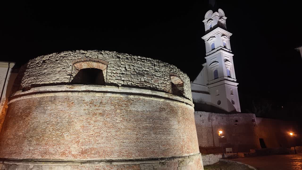 Nighttime view of the Rondella, a historic monument in Sopron, Hungary. The brick structure is beautifully illuminated against the dark sky, with the church tower standing tall in the background.