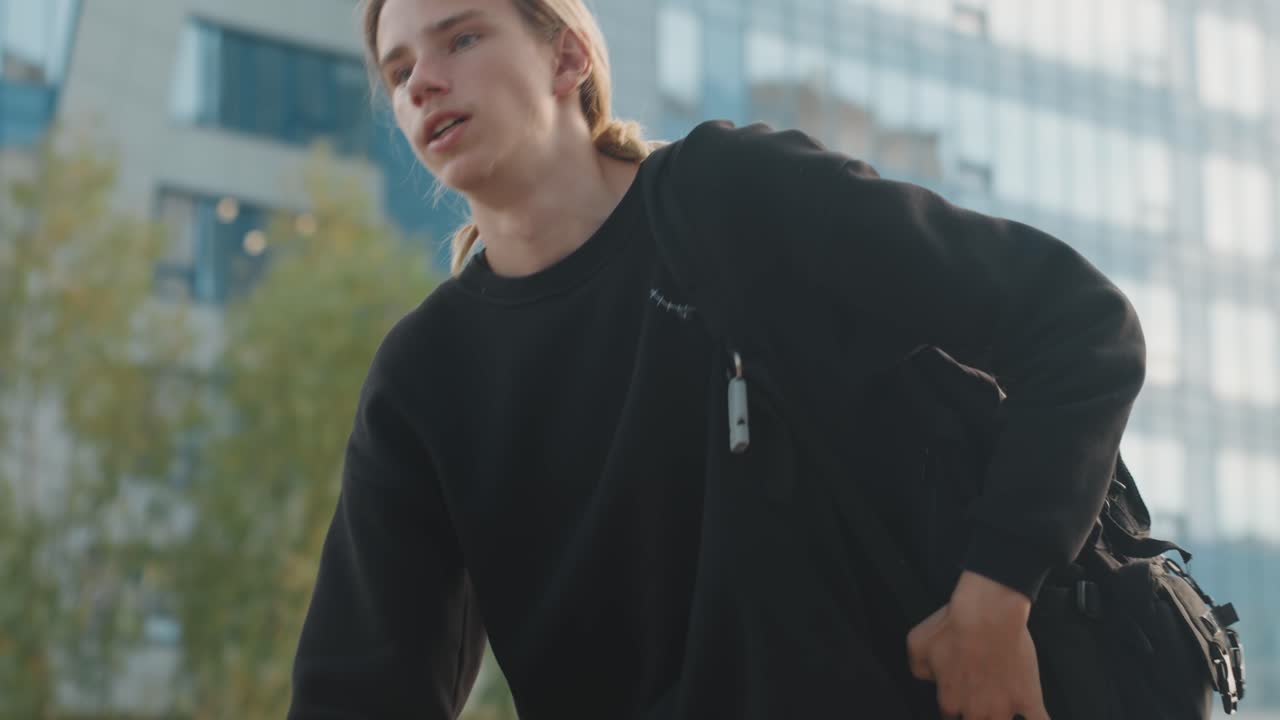 Young man walking with backpack through plaza, glass office towers in background, confident stride and shoulder sling, scooter parked nearby, late afternoon sunlight, urban professional heading