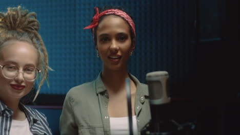 Close-Up-Of-The-Two-Charming-And-Cheerful-Young-Female-Singers-Finishing-Song-Recording-At-The-Studio,-Taking-Off-Headphones-And-Smiling-To-The-Camera