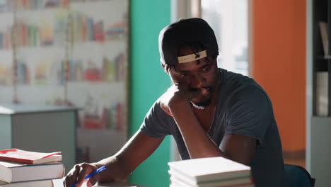 Young man wearing cap sits in library at table by books and notes, studying with pen in hand, resting chin thoughtfully, bright light and wall and shelves behind creating calm contemplative mood
