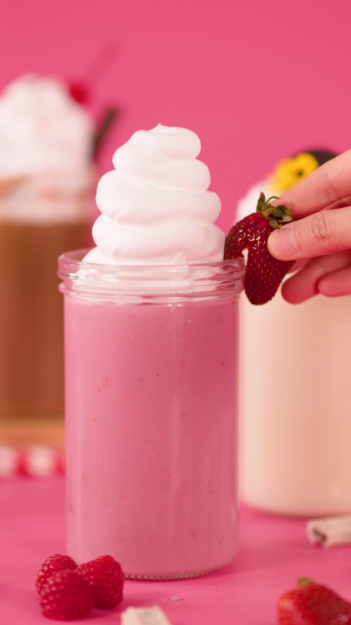 Una mano aggiunge una guarnizione di fragola a un milkshake sormontato da panna montata, sfondo rosa vibrante, illuminazione brillante da studio.