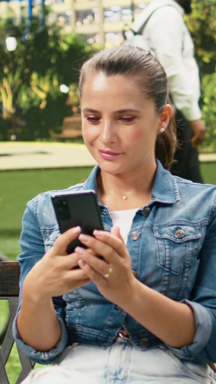 Video verticale: donna sorridente usa lo smartphone mentre si rilassa su una panca di legno.