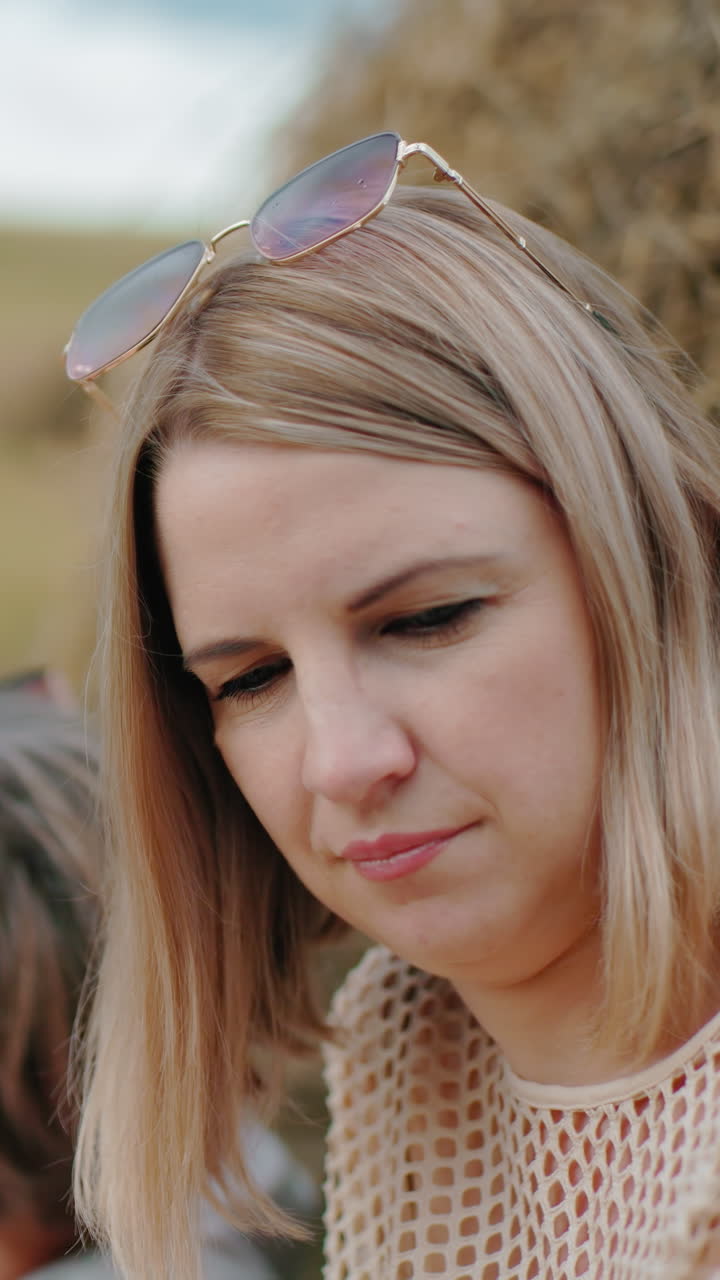 задумчивая женщина с солнцезащитными очками на голове смотрит вниз в спокойной сельской обстановке, другая женщина сидит рядом, с размытой собакой на заднем плане, рядом с большим пакетом сена на обширной открытой сельскохозяйственной земле