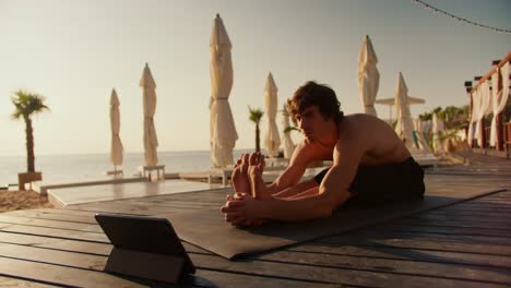The-guy-does-the-stretch-following-the-video-on-the-tablet.-Online-stretching-and-yoga-classes.-Sunny-beach-in-the-morning