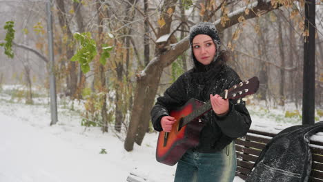 young woman in black hoodie playing guitar outdoors in snowy park, standing near snow-covered bench, surrounded by frosty trees and softly falling snow