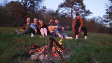 pieces of wood burning slowly in the campfire. company of youngsters sitting on the grass, playing guitar and singing. blurred background.