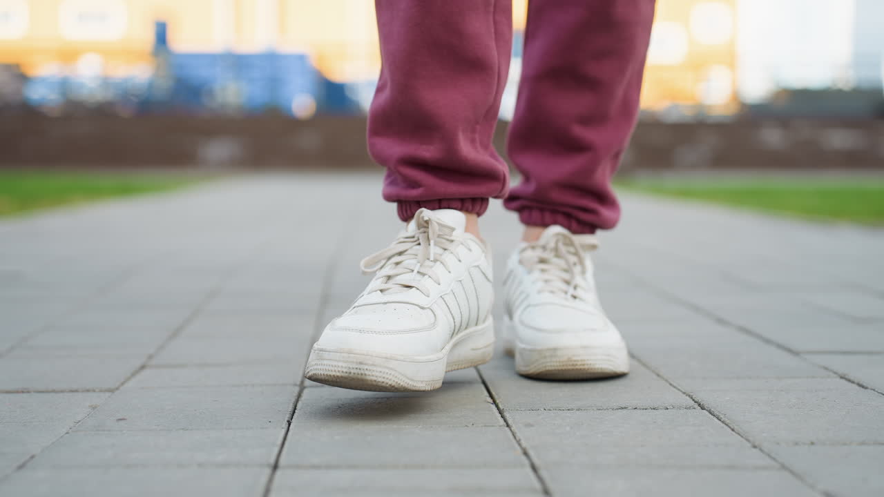 Close up leg view of female jogger wearing white sneakers and burgundy jogger trousers stepping forward on tiled pavement in urban street scene capturing dynamic motion and footwear texture detail