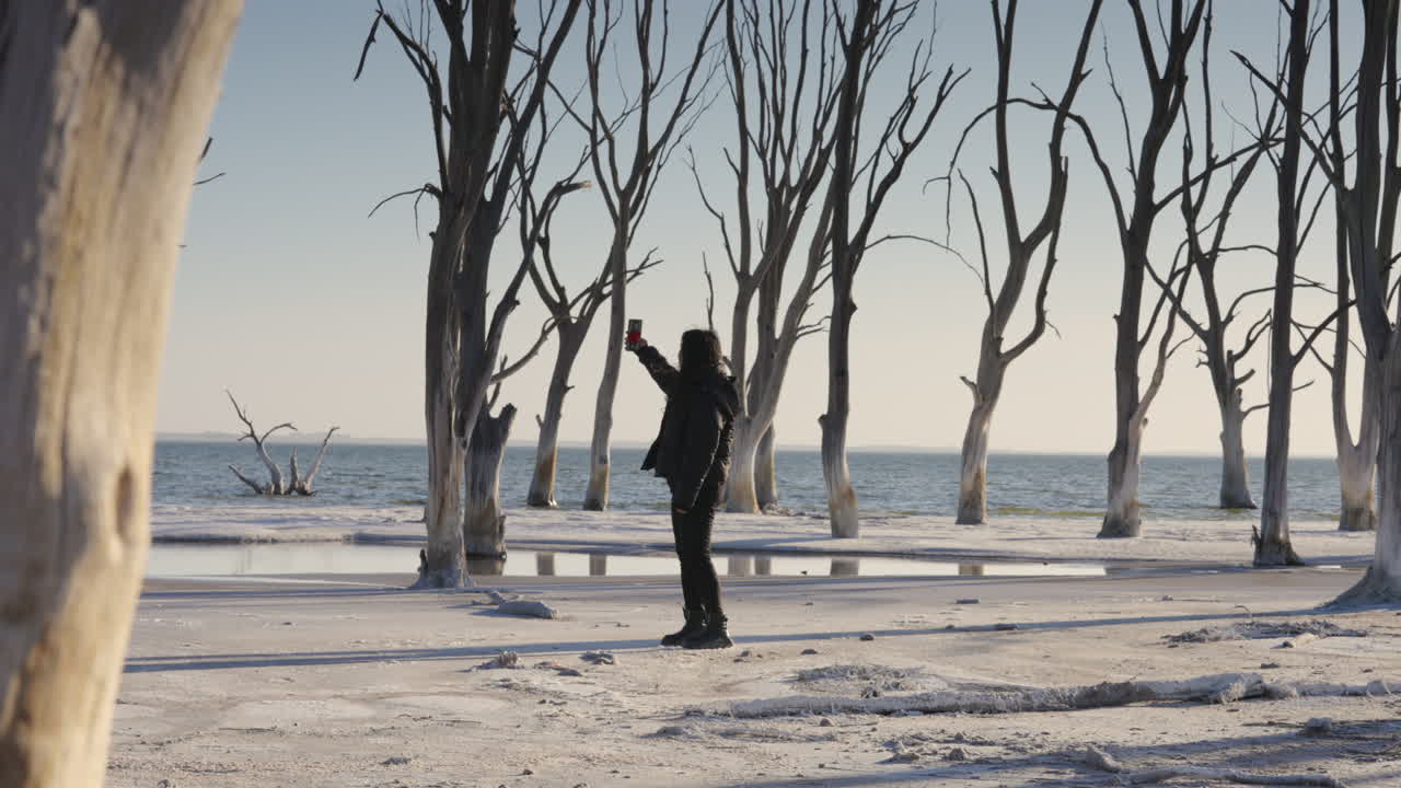 Donna che Cerca Segnale del Telefono Cellulare in un Paesaggio Costiero Brullo con Alberi Secchi e Mare sullo Sfondo