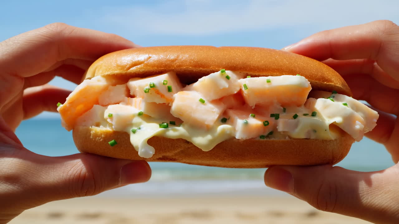 Roll de langosta sostenido sobre la playa, manos iluminadas por el sol presentando ensalada cremosa de langosta en pan suave con cebollino, océano.