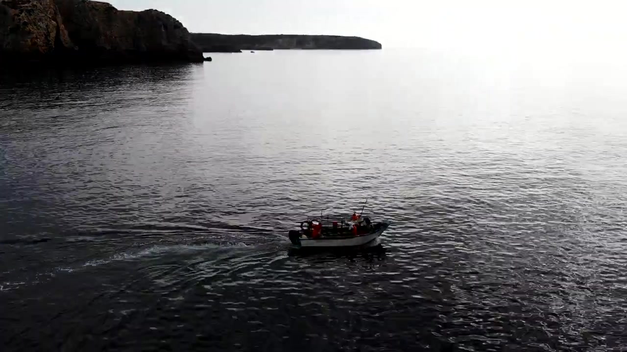 Piccola motovedetta vicino a scogliere, equipaggio che opera con poca luce e scia stabile, scogliere stagliate contro un cielo pallido.