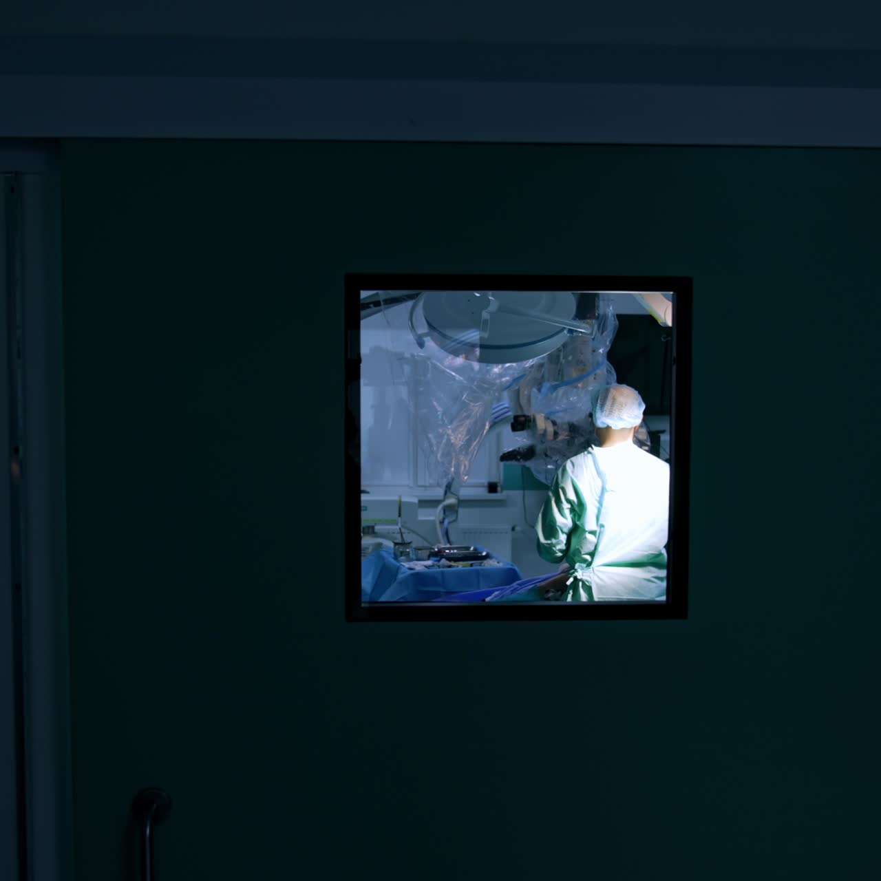 Footage through the square window in a surgery room door. Busy doctors are performing surgery at operational table. Screen in the operational theatre showing the procedure