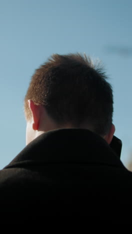 back view of man in black coat engaged in phone call outdoors, shaking head in displeasure while reacting to intense conversation, with urban background including modern buildings, greenery