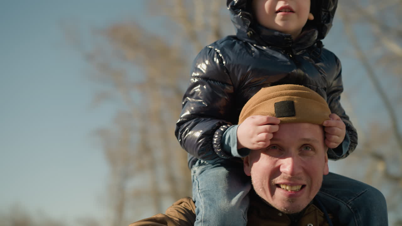 A father carries his young son on shoulders while pointing out something interesting, covering his father's eyes with hands and adjusting beanie