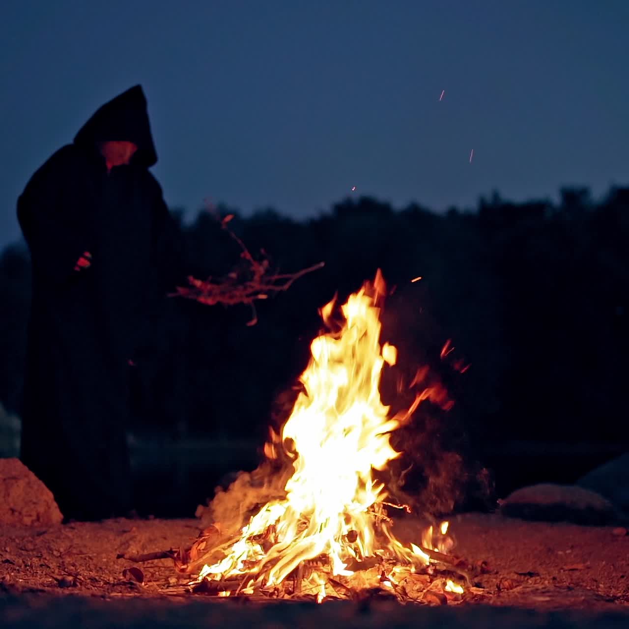 Witch in black robe doing ritual near the bonfire. Vivid fire and sorcerer. Mysterious dark figure conjuring outdoors. Halloween.