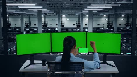 Back View Of Happy Asian Woman Looking At Mobile Phone Green Screen And Celebrating While Working With Multiple Computer Monitor In Data Center