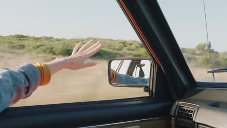 Frau-Hält-Die-Hand-Aus-Dem-Autofenster-Und-Spürt,-Wie-Der-Wind-Durch-Die-Finger-Weht,-Während-Sie-Auf-Dem-Land-Fährt-Und-Im-Sommerurlaub-Unterwegs-Ist-Und-Die-Freiheit-Auf-Der-Straße-Genießt