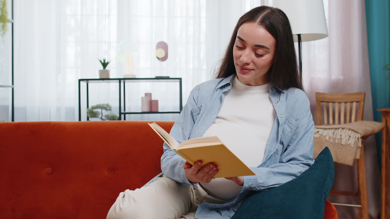 Pregnant caucasian woman reading book advice child care guide feeling relax on cozy couch at home