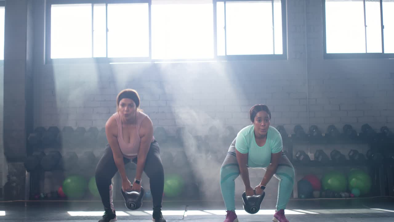 Diverse partner di allenamento femminili che eseguono squat-to-press con kettlebell in palestra, spinta dell'anca per la forza