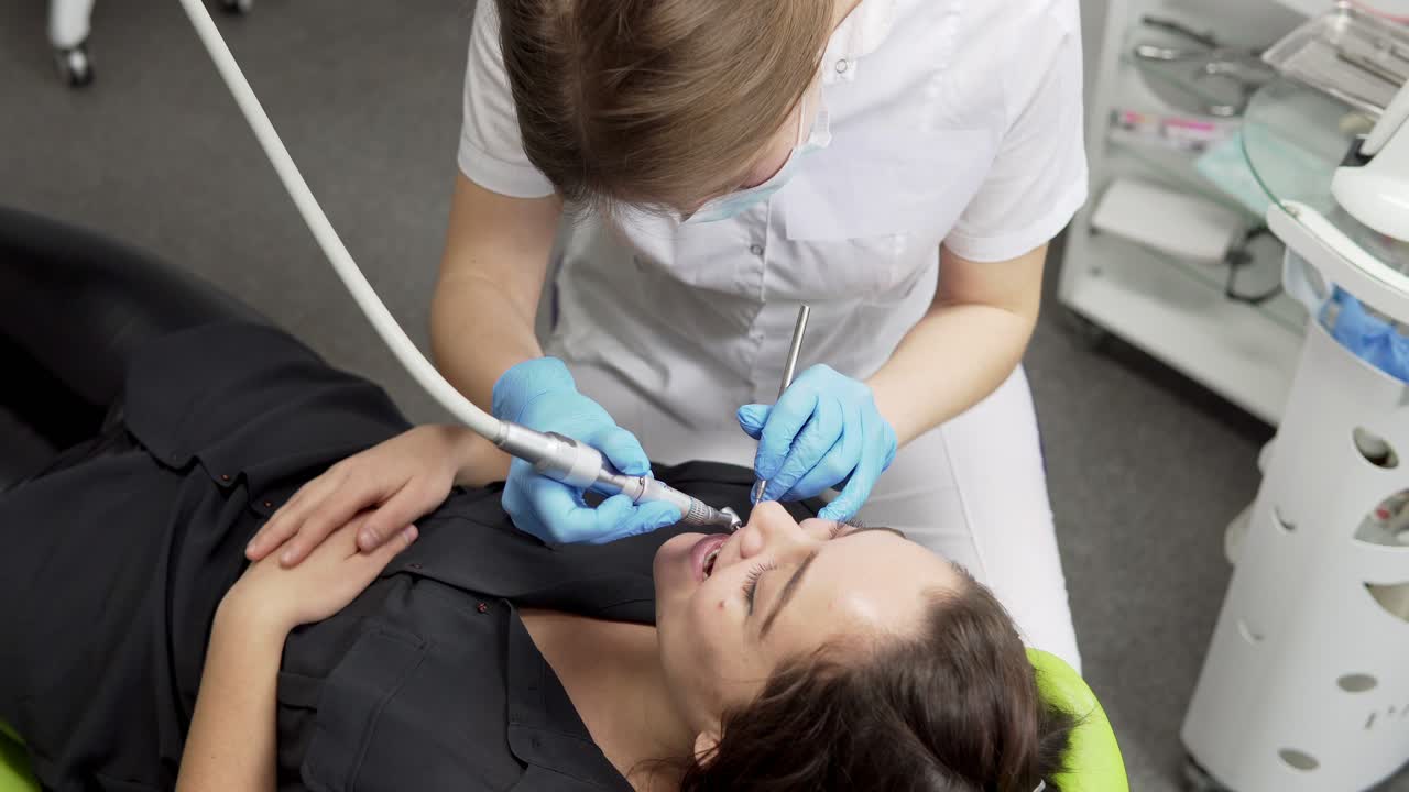 Young female dentist in gloves and mask drilling patient's teeth in clinic