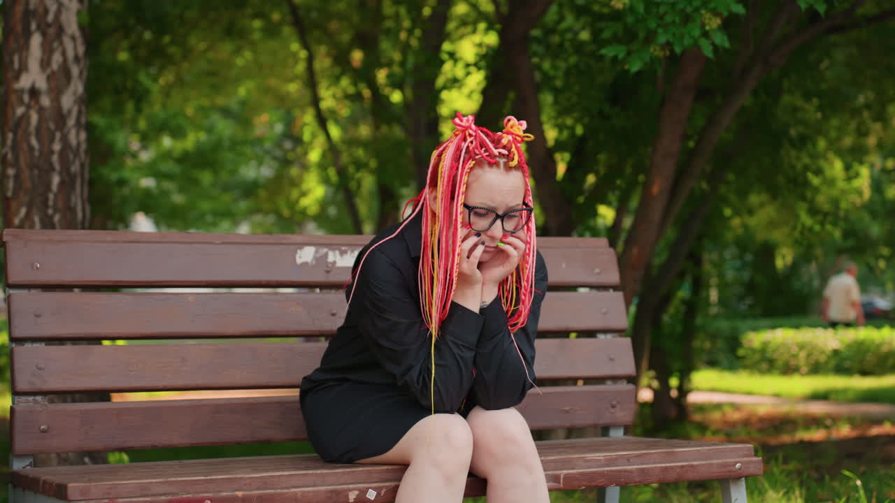 알록달록한 드레드락 머리를 하고 검은색 드레스를 입은 젊은 백인 여성이 벤치에 앉아 두 손으로 얼굴을 감싸고 있어요. 한적한 낮 시간의 공원, 자작나무, 부드러운 햇빛 아래에서 슬픔을 표현하는 감정적인 인물 사진이에요.