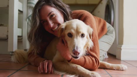 smiling caucasian woman kissing and cuddling her pet dog sitting on floor at home